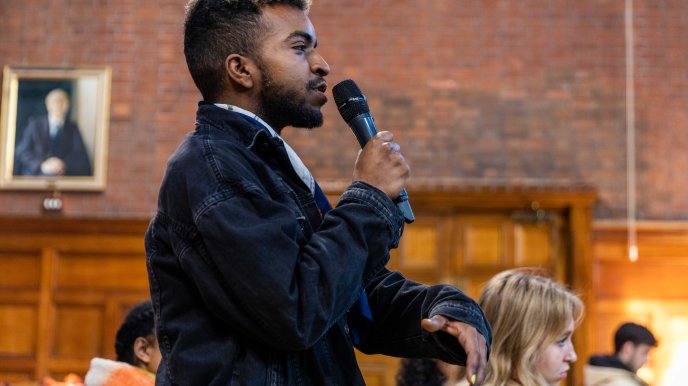 A young Black man with a beard stands with a microphone to ask a question
