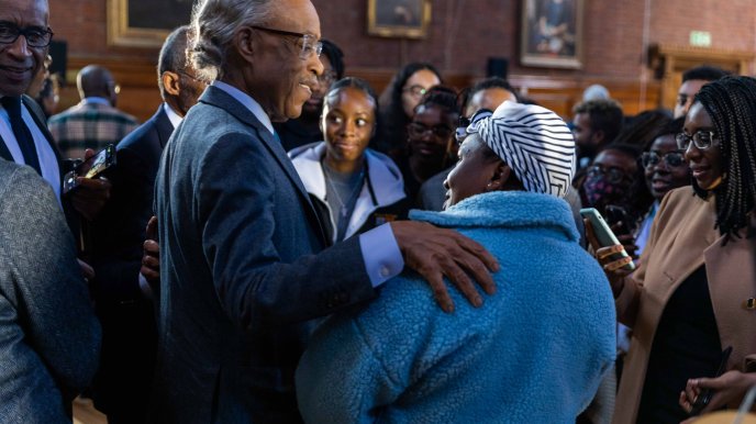 Rev Sharpton stands with his hand on the shoulder of a shorter Black woman wearing a blue winter coat